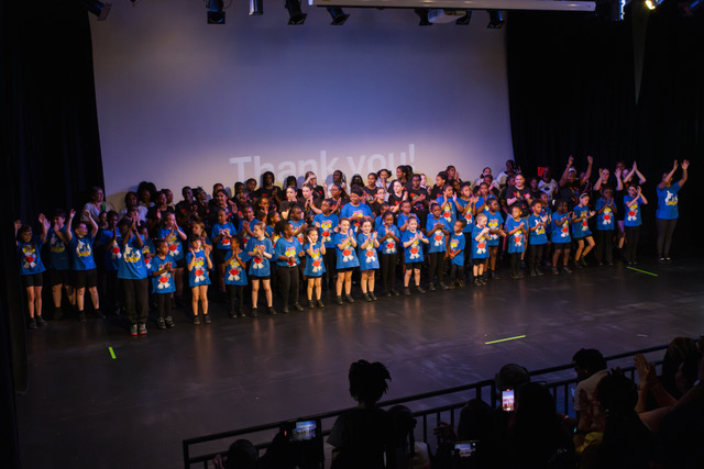 A large group of young people stand in a stage wearing matching blue t-shirts